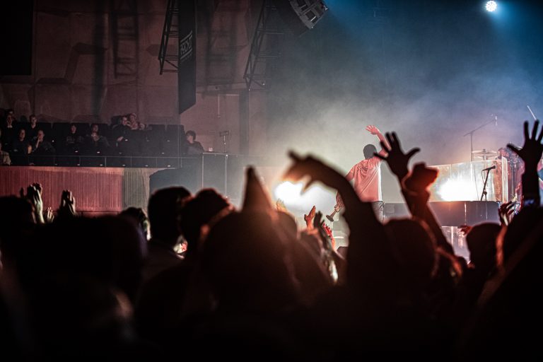 Een menigte met opgestoken handen staat voor een podium tijdens een liveoptreden in een sfeervolle, verlichte omgeving.
