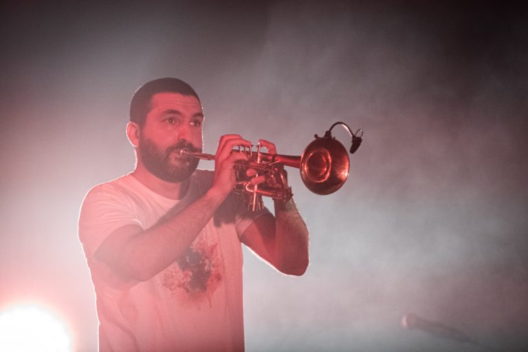 Een man speelt op een trompet terwijl hij op het podium staat, omringd door rook en sfeerlicht.