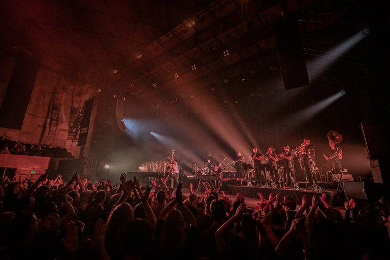 Een live concert met een enthousiaste menigte die applaudisseert, terwijl een band op het podium speelt onder fel licht.