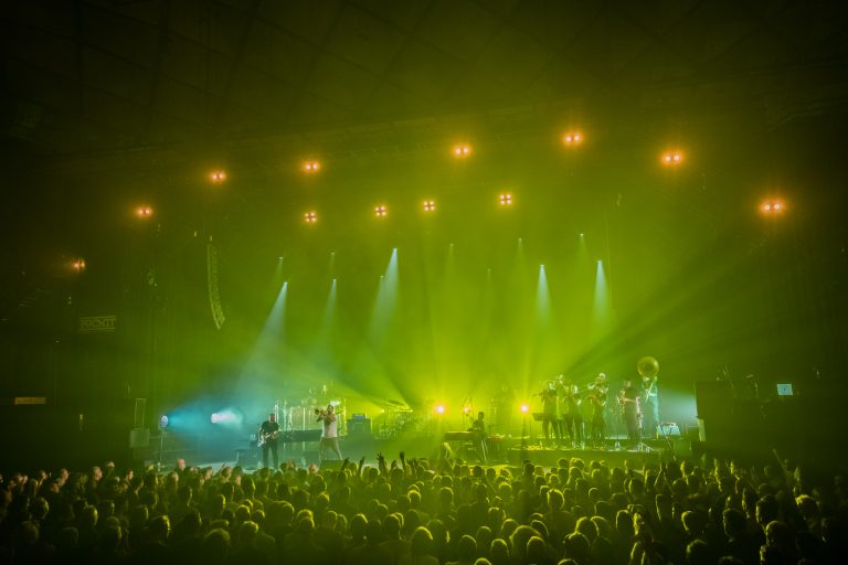 Een druk concert met een energievolle band, verlicht door groene en blauwe lichten.