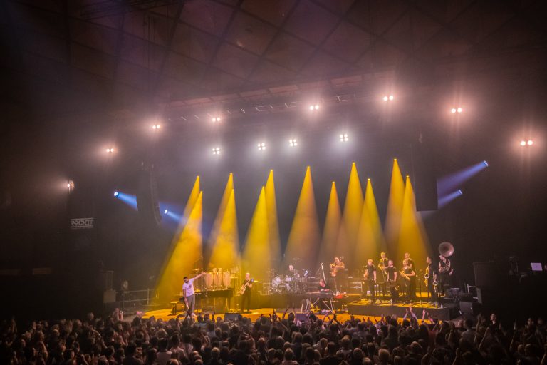 Een live muziekoptreden met een grote band op het podium, verlicht door gele lichten, voor een enthousiast publiek.