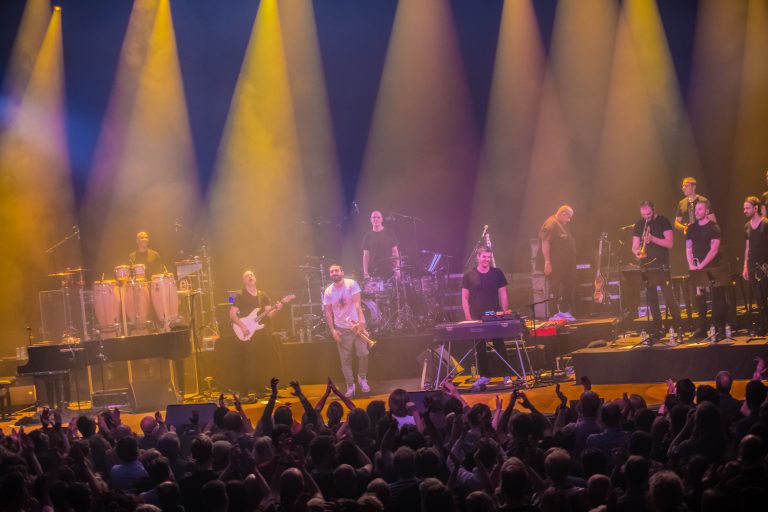 Een live band staat op het podium met kleurrijke lichten, terwijl het publiek enthousiast reageert.