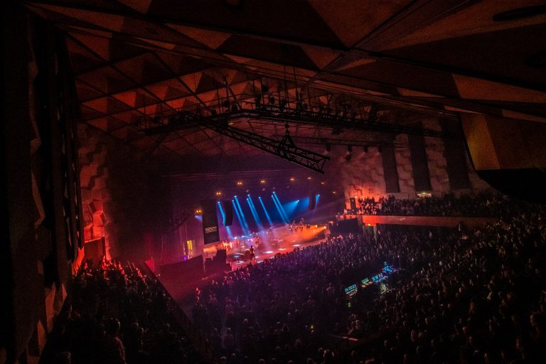 Een druk bezochte concertzaal met kleurrijke lichten en een groot publiek dat geniet van de live muziek.
