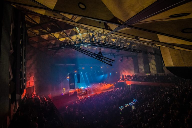 Een druk concert met kleurrijke verlichting en een grote menigte in een indoorsetting.