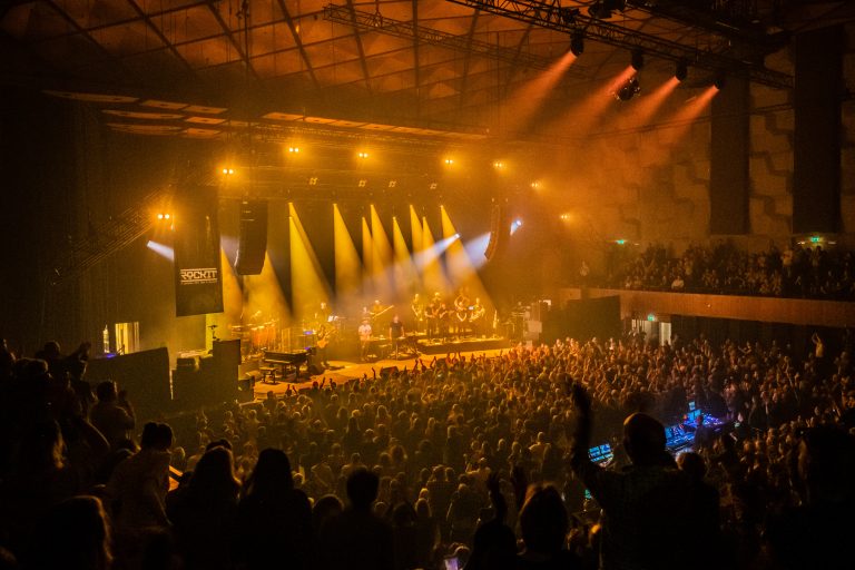 Een druk concert met een groot publiek en heldere lichteffecten op het podium.