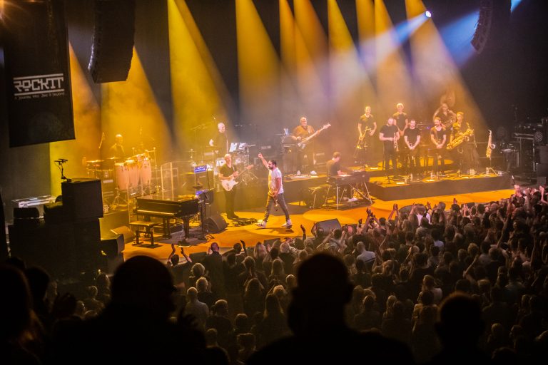 Een publiek geniet van een live optreden op een podium verlicht door gele en blauwe lichten.