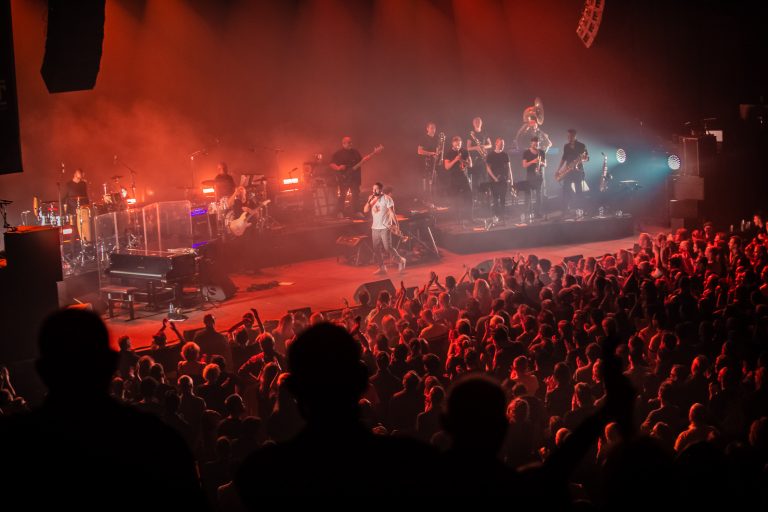 Een live concert met een grote menigte en een band op het podium, omringd door kleurrijke verlichting.