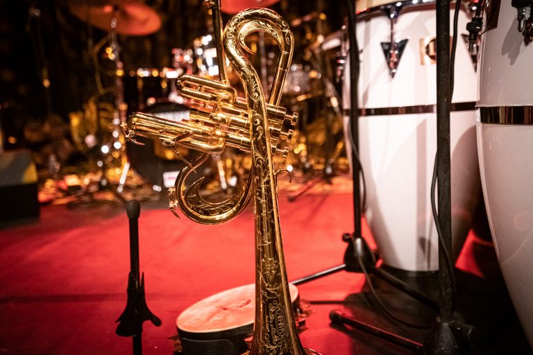 Een gouden muziekinstrument, mogelijk een trompet, staat op een rode vloer voor andere muziekinstrumenten.