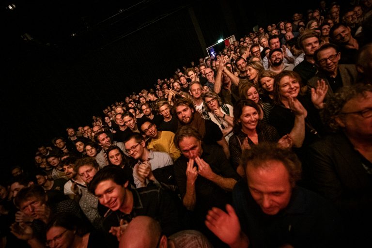 Een grote menigte mensen die enthousiast applauss en geniet van een optreden in een donkere zaal.