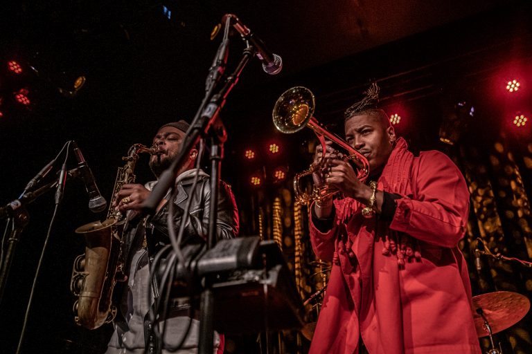 Twee muzikanten op het podium spelen respectievelijk saxofoon en trompet in een sfeervolle, verlichte setting.
