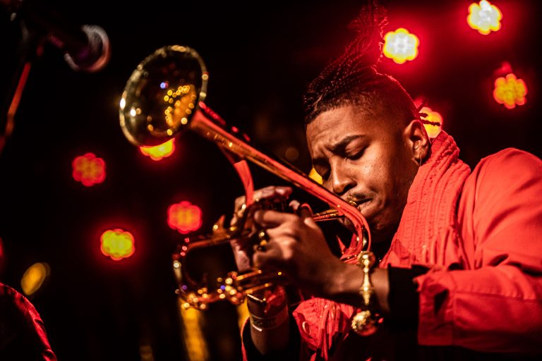 Een muzikant speelt gepassioneerd op een trompet met warmrode lichten op de achtergrond.