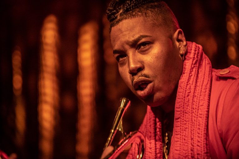 Een zanger in een felgekleurde outfit, die intens in de camera kijkt terwijl hij een microfoon vasthoudt.