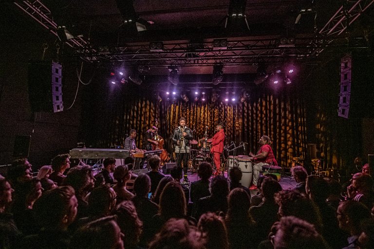 Een liveband treedt op voor een publiek in een sfeervolle concertzaal met paarse verlichting.