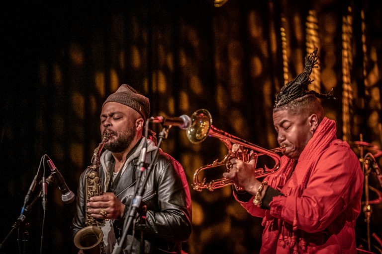 Twee muzikanten spelen op het podium, één met een saxofoon en de ander met een trompet.