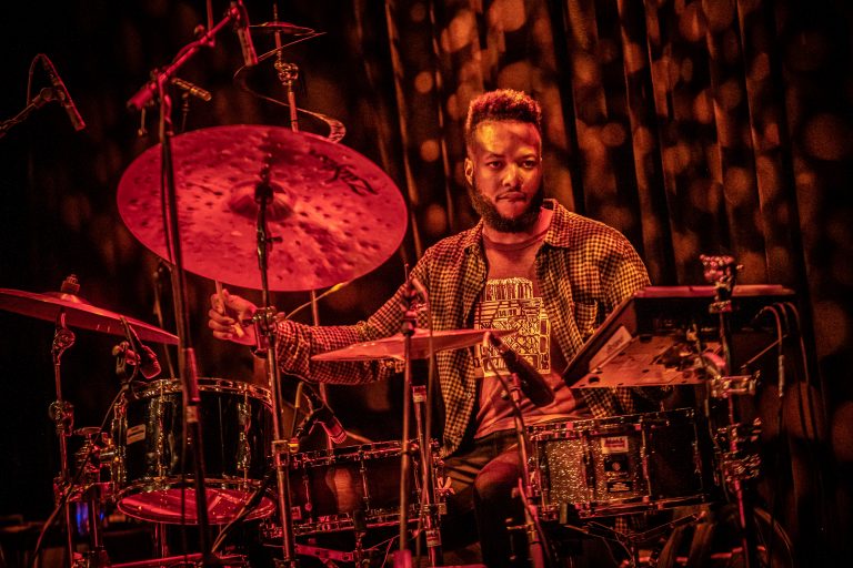 Een drummer speelt op zijn drumstel, omringd door een warme rode gloed.