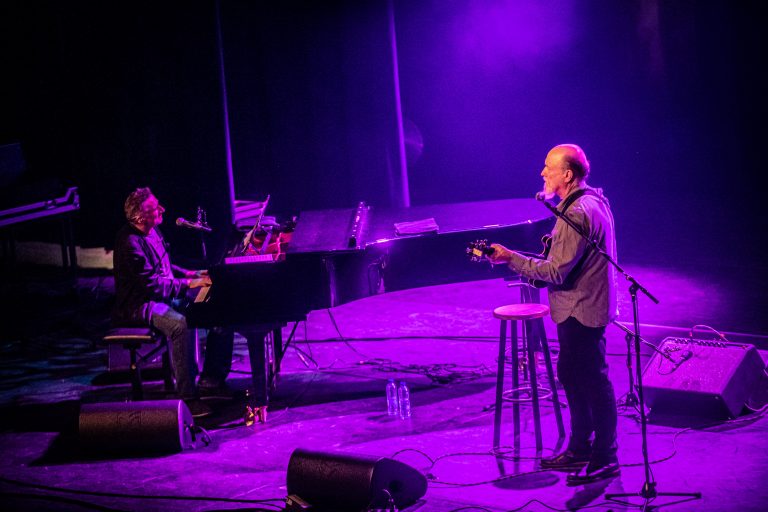 Twee muzikanten treden op het podium op, met een piano aan de linkerkant en één van hen die een gitaar speelt.