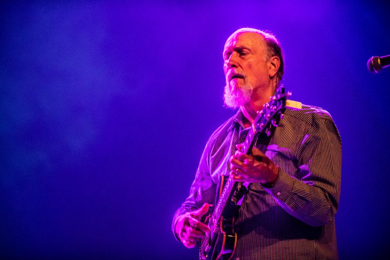 Een muzikant speelt gitaar op een podium met paarse verlichting.