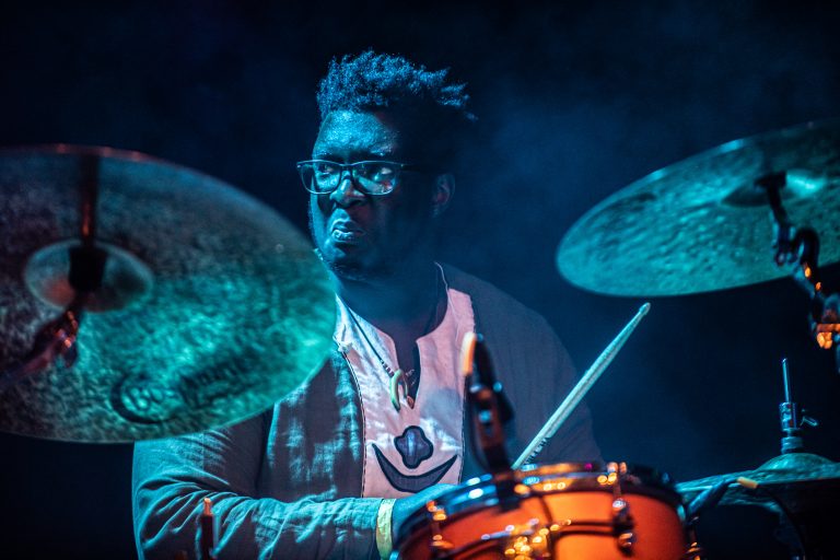 Een drummer speelt intensief op zijn instrument terwijl er blauwe verlichting op hem schijnt.