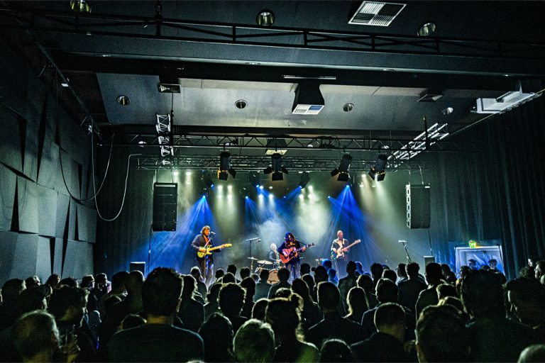Een live band speelt op een podium voor een publiek in een sfeervolle concertzaal met kleurrijke verlichting.