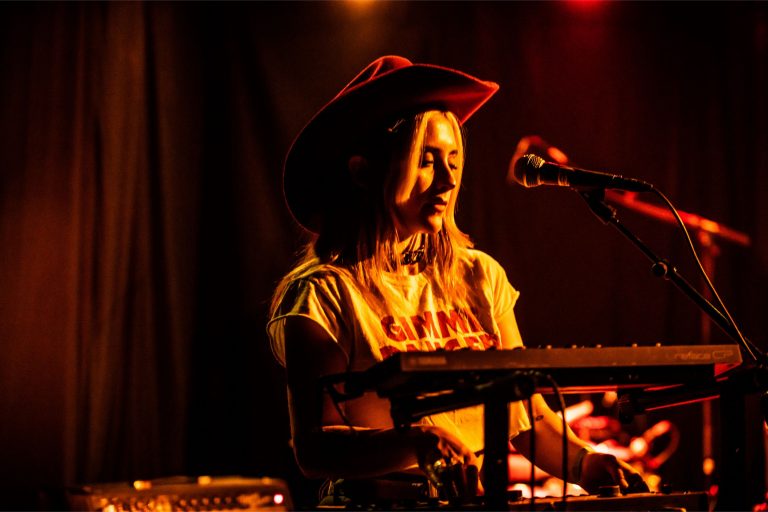 Een muzikant in een cowboyhoed die achter een keyboard staat, belicht door warme lichten.