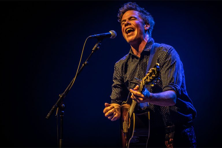 Een vrolijk uitziende muzikant zingt en speelt gitaar op een podium met een blauwe achtergrond.