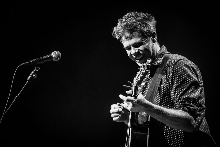 Een man met een gitaar lacht terwijl hij op het podium speelt.