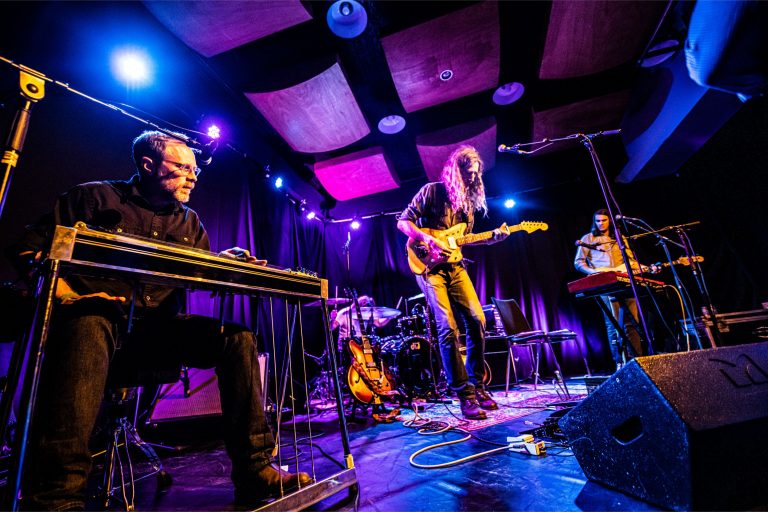 Een live muziekopvoering met drie muzikanten op het podium, omringd door kleurrijke verlichting.