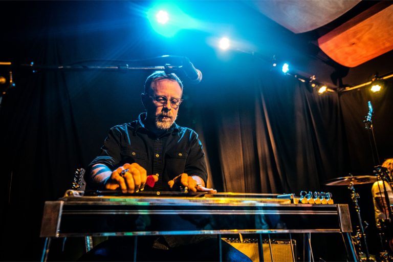 Een muzikant speelt een stalen gitaar op een podium, verlicht door blauwe en gele lichten.