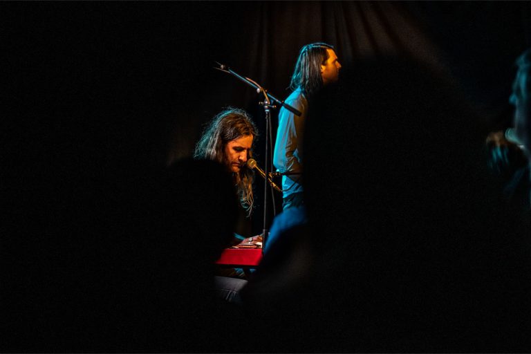 Twee muzikanten zijn te zien, waarbij de ene aan een keyboard speelt en de andere zingt, omringd door schaduwen van het publiek.