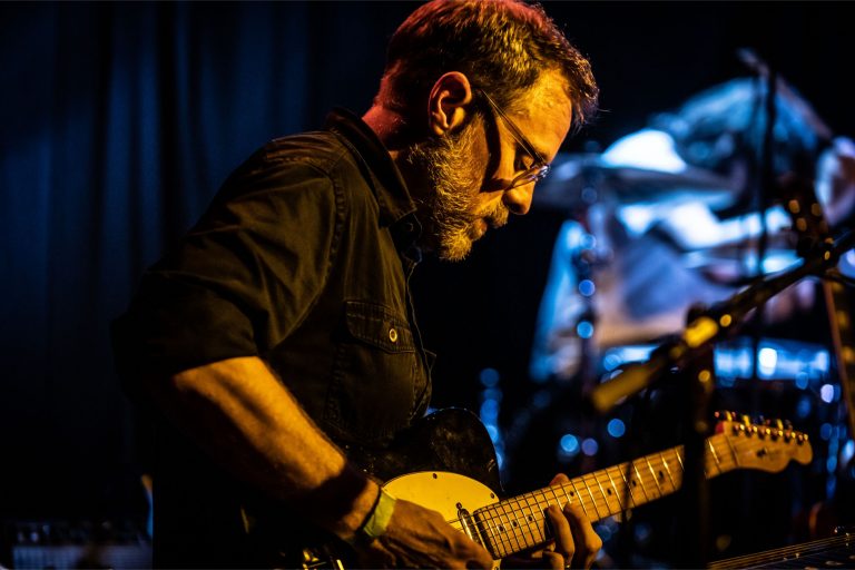 Een muzikant speelt gitaar op een podium met een donkere achtergrond en sfeervolle verlichting.
