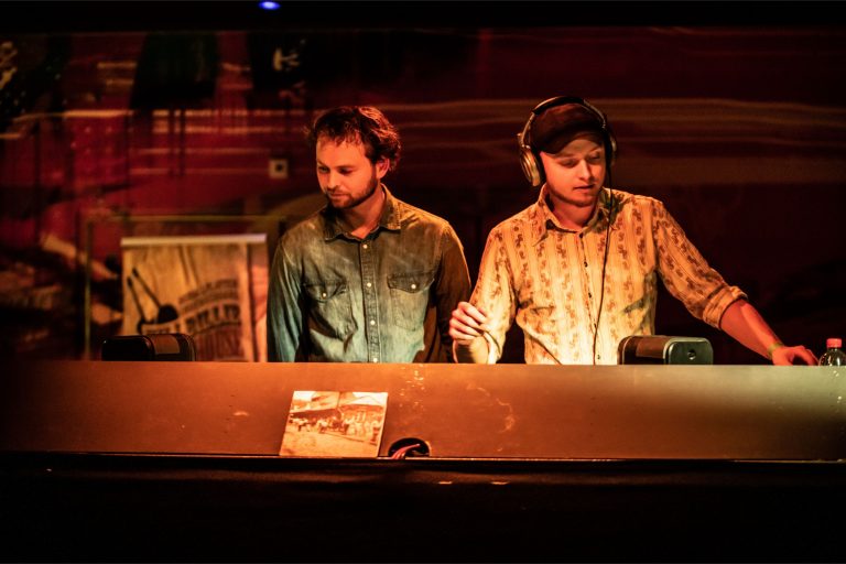 Twee mannen staan achter een DJ-tafel in een sfeervolle, verlichte omgeving.