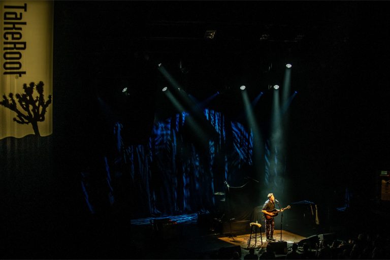 Een muzikant speelt gitaar op een podium, omringd door kleurrijke verlichting.