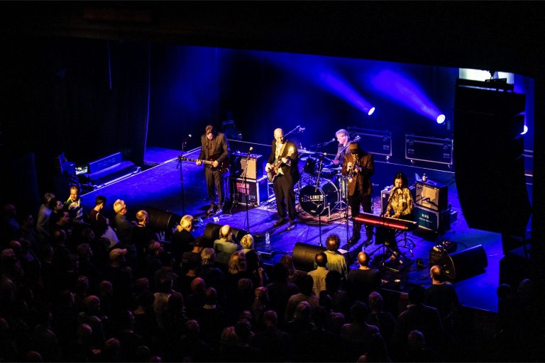 Een live band speelt op een podium verlicht met paarse lichten voor een enthousiast publiek.