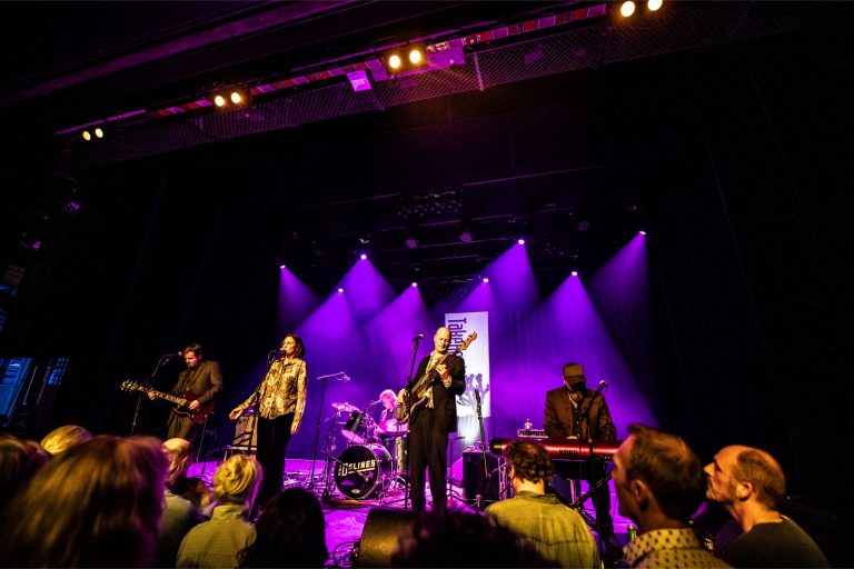 Een band speelt op een podium, omringd door paarse verlichting, met een publiek dat naar de voorstelling kijkt.