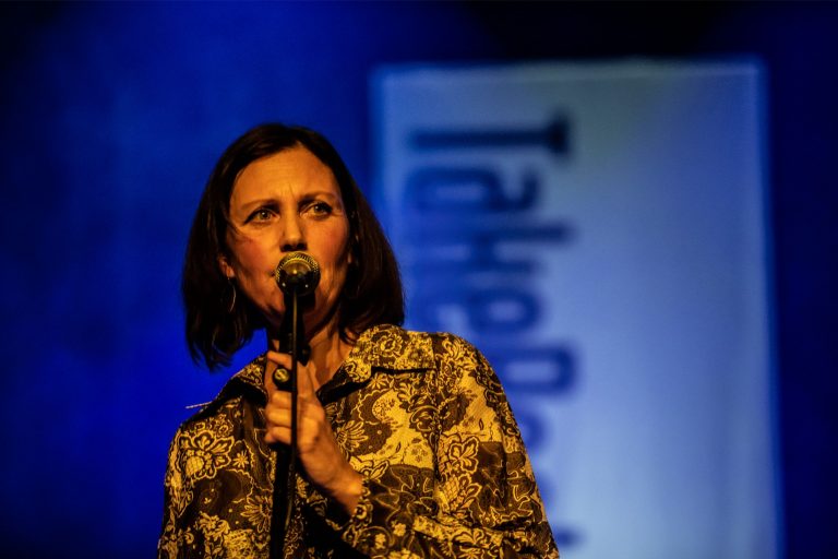 Een vrouw met een microphone, gekleed in een gedurfde blouse, staat op een podium met een blauwachtige achtergrond.