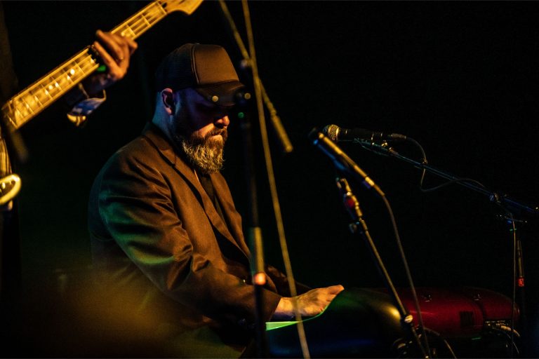 Een muzikant met een hoed en een baard speelt op een toetsenbord, met een microfoon en een gitaar zichtbaar op de achtergrond.