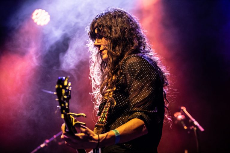 Een muzikant met lang, golvend haar speelt gitaar op een podium met kleurrijke belichting.