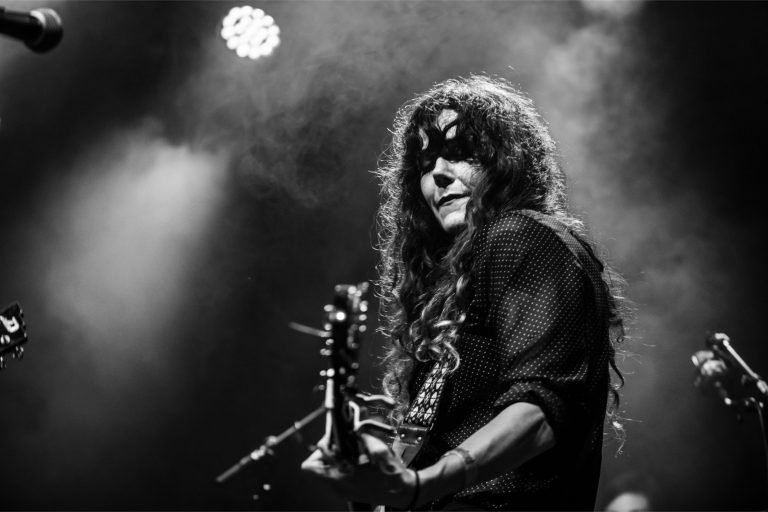 Een muzikant met lang haar en een masker speelt gitaar op een podium, omgeven door sfeervolle verlichting.