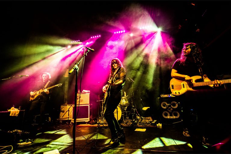 Een rockband speelt live op het podium, omringd door kleurrijke lichteffecten.