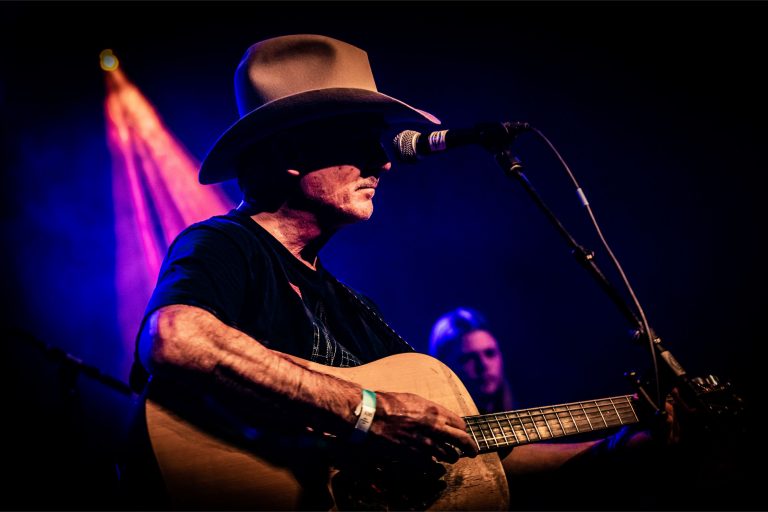 Een muzikant met een hoed speelt gitaar en zingt in een live-muzieksetting, verlicht door kleurrijke lichten.