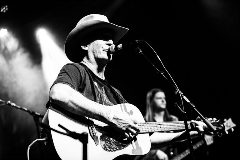 Een muzikant met een hoed speelt gitaar en zingt in een microfoon op het podium, met een andere muzikant op de achtergrond.