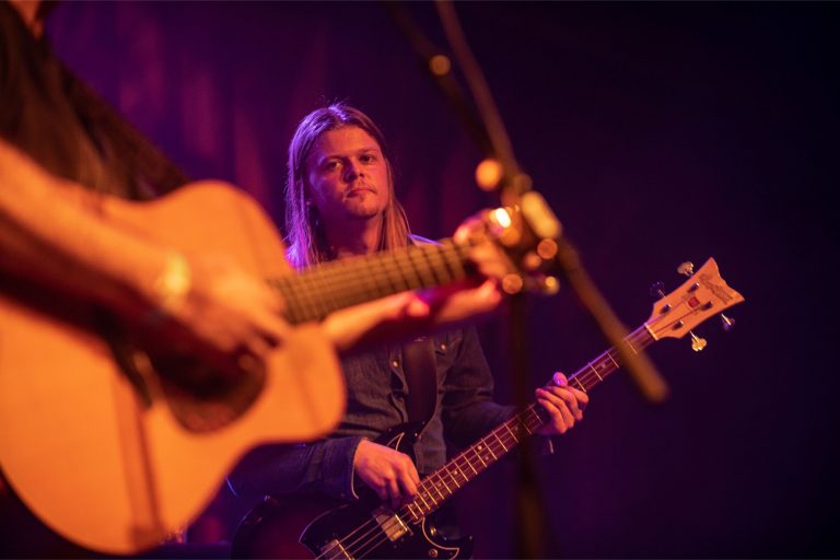 Een muzikant speelt gitaar op het podium, terwijl een andere muzikant met lang haar de basgitaar bespeelt op de achtergrond.