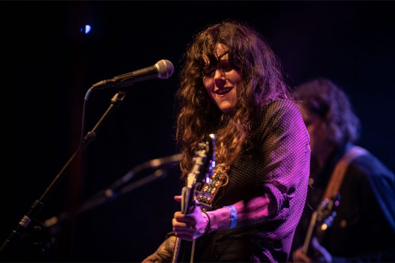 Een muzikante met een gitaar staat op een podium en zingt in een sfeervolle lichtsetting.