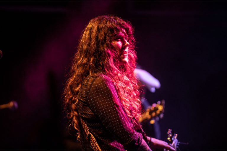 Een vrouw met lang, krullend haar zingt op een podium terwijl ze naar de zijkant kijkt, verlicht door kleurrijke lichten.