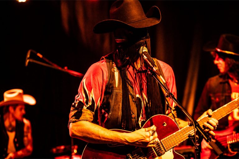 Een muzikant met een hoed speelt gitaar en zingt op een podium met een warme, sfeervolle achtergrondverlichting.