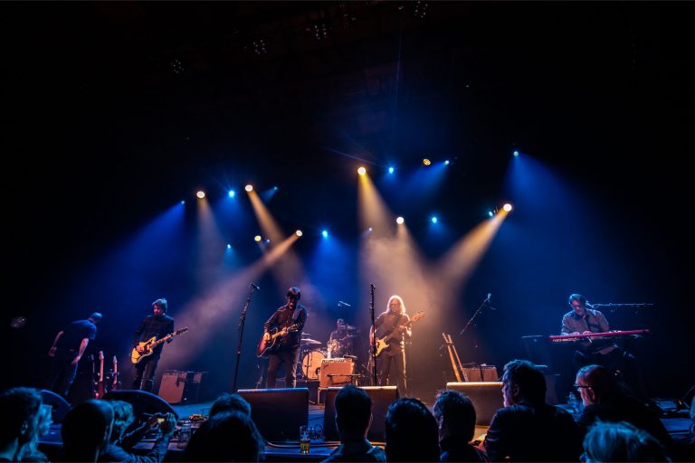 Een band staat op een podium met kleurrijke verlichting terwijl ze een live optreden geven voor een publiek.