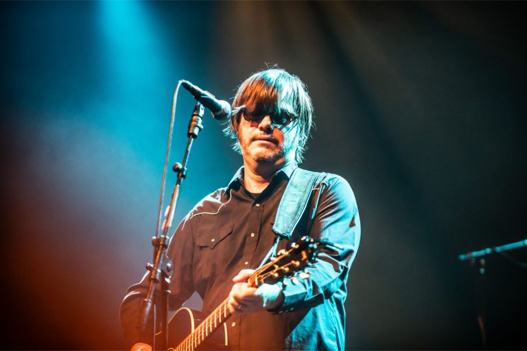 Een muzikant met een gitaar staat op een podium onder kleurrijke lichten.