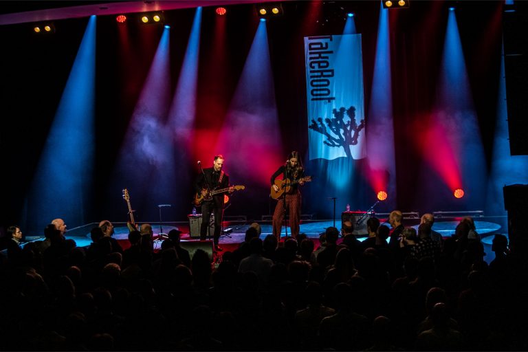 Twee muzikanten staan op een podium met gekleurde lichtstralen en een banner op de achtergrond.
