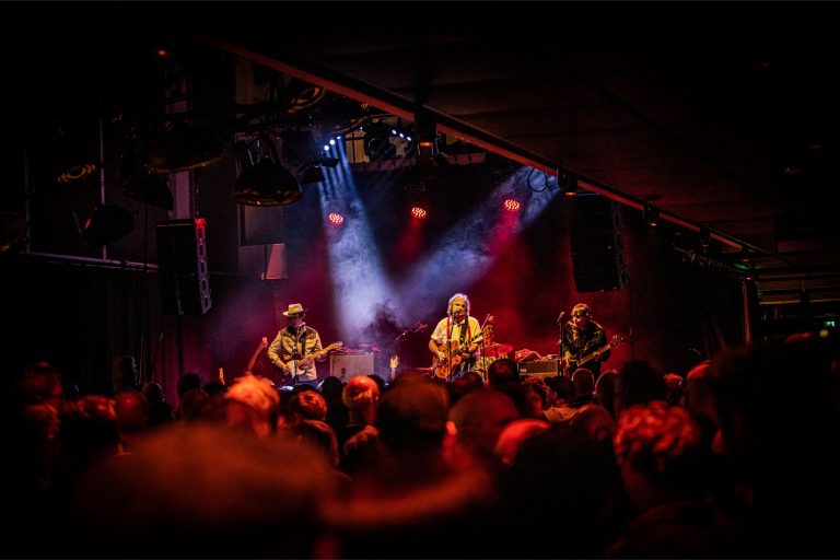 Een band speelt live muziek op een podium, omringd door een menigte met kleurrijke lichten.