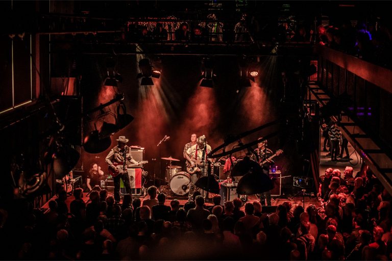 Een band speelt op een podium voor een groot publiek, omgeven door sfeervolle rode verlichting.